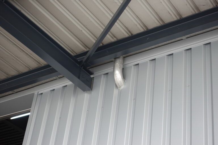 Aussenfassade des Gebäudes aus grauem Metall mit Wellblechverkleidung, Stahlträgern und einem weissen Kunststoff-Abflussrohr.