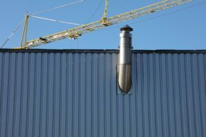 Ein glänzendes, zylindrisches Metallkaminrohr an einer Wellblechwand; im Hintergrund ein Kran.