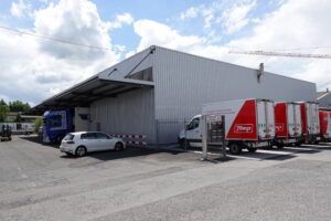 Lagerhalle mit Lieferwagen und einem davor geparkten Auto. Graues Gebäude, blaue und rote Fahrzeuge.