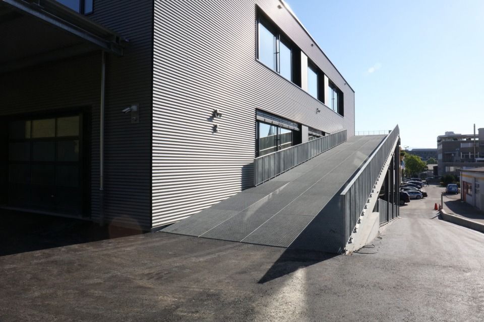 Aussenansicht eines modernen Gebäudes mit einer langen Rampe, die zu einem Eingang im zweiten Stock führt; sonniger Tag.