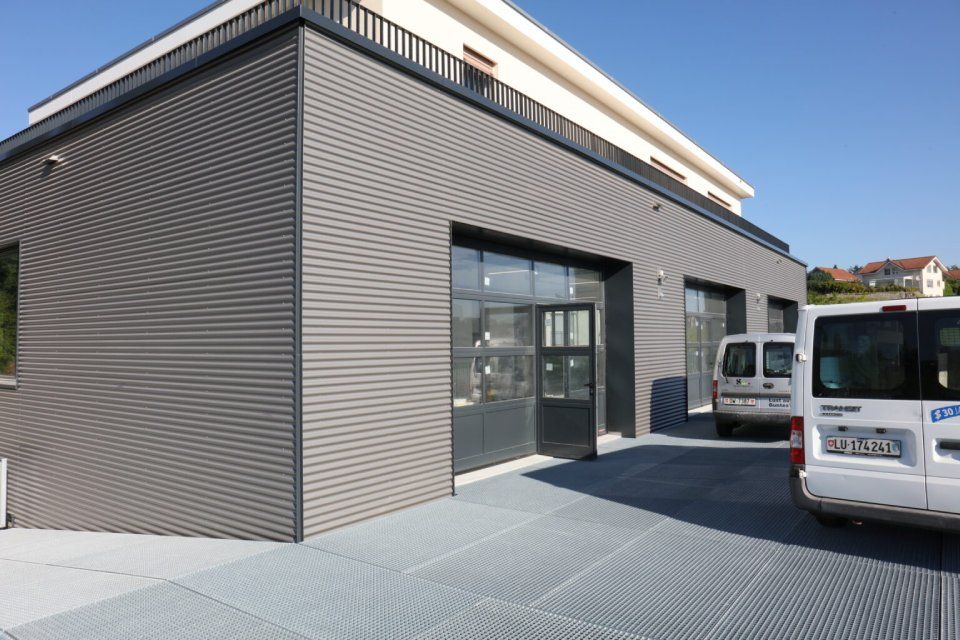 Graues Gebäude mit grossem Garagentor, zwei Lieferwagen stehen draussen auf einer Metallgitterfläche.