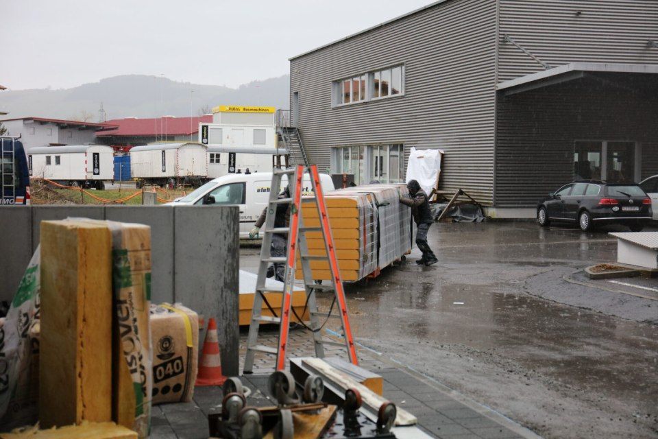 Ein Mann schiebt einen Stapel Dämmplatten auf einem Anhänger vor einem Gebäude. Szene im Freien bei Nässe.