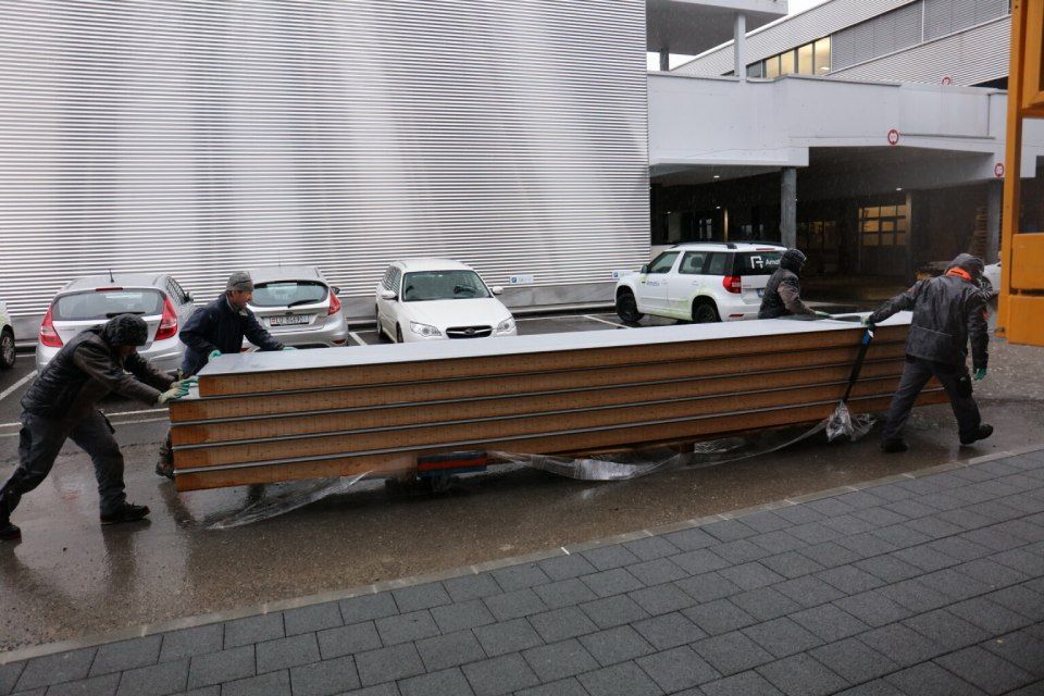 Drei Arbeiter schieben einen grossen Stapel Holzplatten auf einer regnerischen Aussenfläche in der Nähe von geparkten Autos und einem Gebäude.