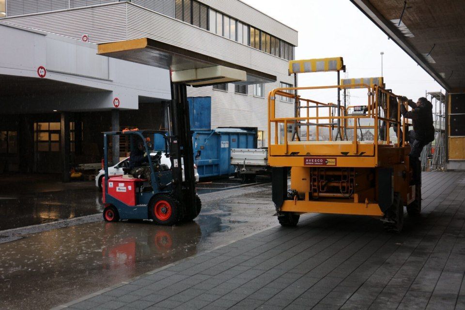 Ein Gabelstapler steht neben einer gelben Hebebühne; eine Person steigt im Freien in der Nähe eines Gebäudes darauf.