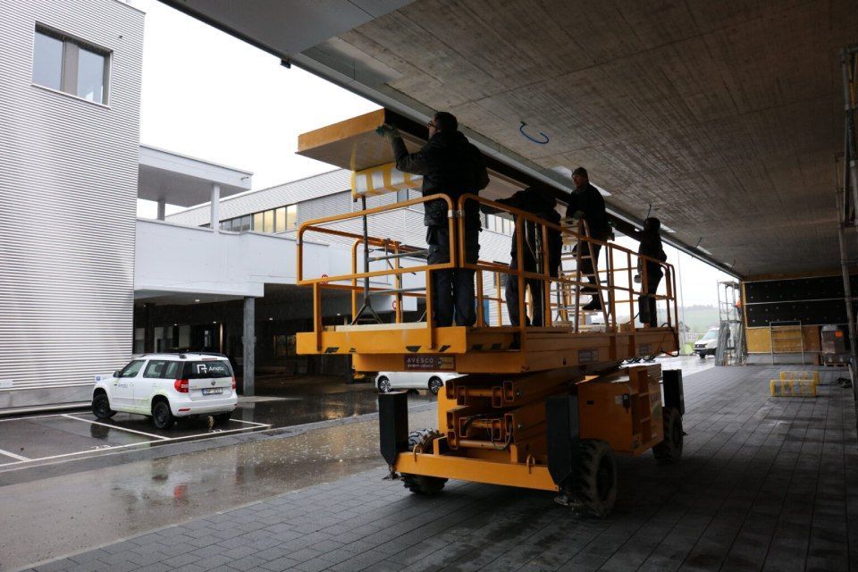 Drei Arbeiter montieren auf einer gelben Hebebühne etwas unter dem Dachvorsprung eines Gebäudes. Ein weisses Auto ist in der Nähe geparkt.