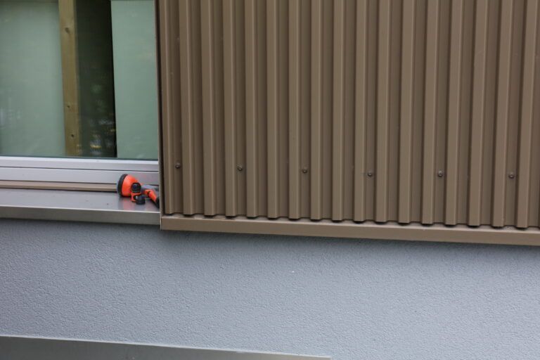 Fenster mit brauner Wellblechverkleidung an einer grauen Aussenwand, orangefarbenes Werkzeug auf der Fensterbank.