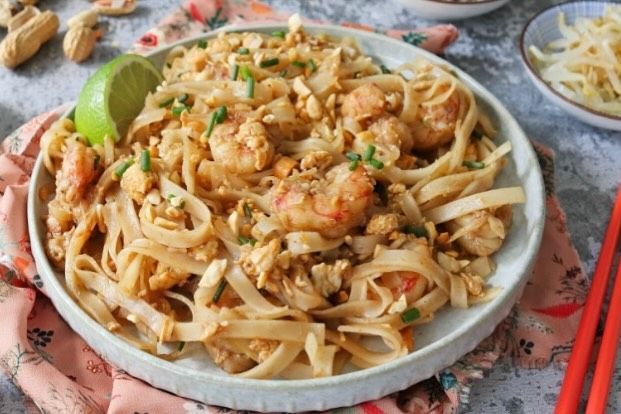 Plato de pasta con fideos y camarones con lima sobre un mantel rosa, con palillos y guarniciones a mano.
