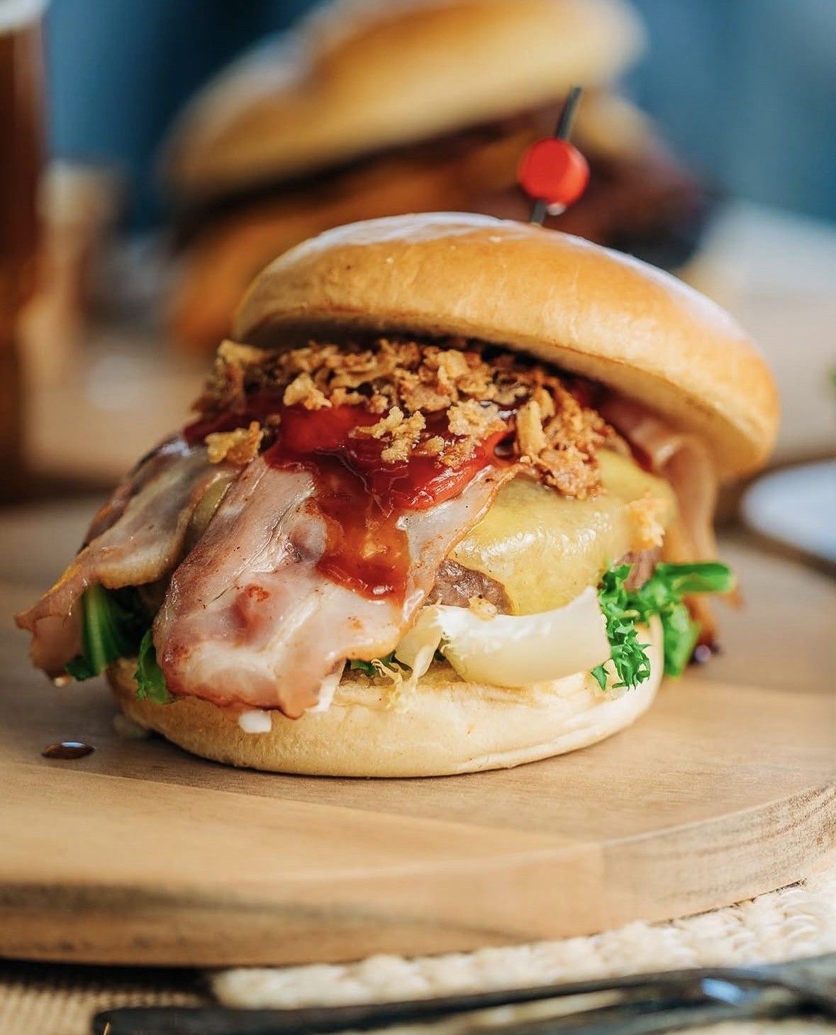 Primer plano de una hamburguesa con queso, tocino, salsa y un tomate cherry sobre una tabla de madera.