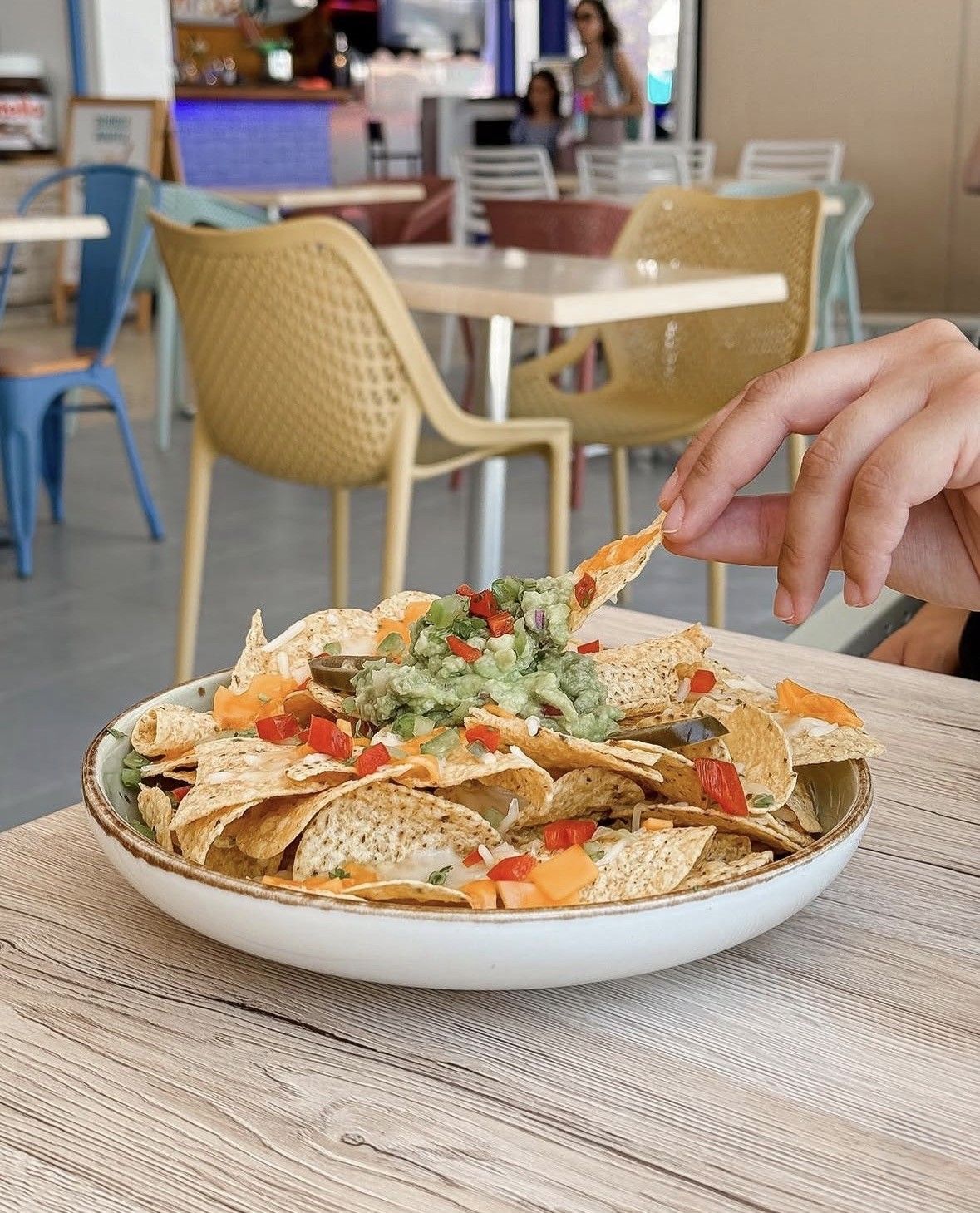 Una mano levanta un nacho de un plato sobre la mesa de una cafetería, con el restaurante de fondo borroso.