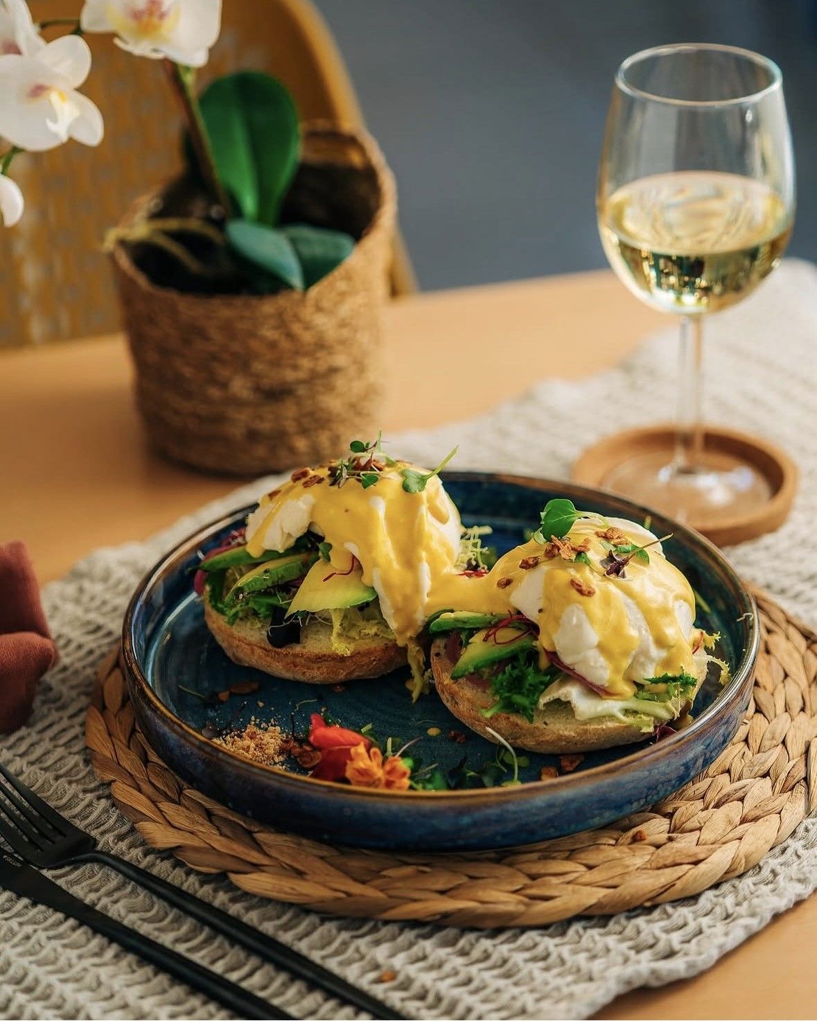 Tostada de aguacate en un plato azul con huevos escalfados, sobre un mantel individual tejido junto a una copa de vino 