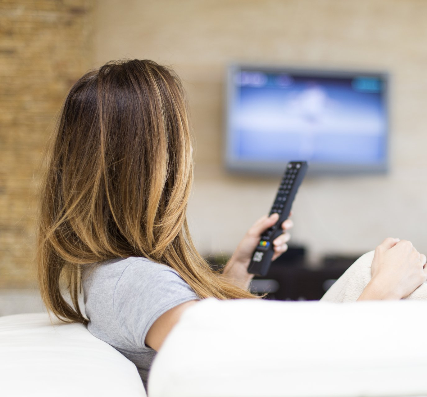 Femme aux longs cheveux assise sur un canapé, regardant la télévision, tenant une télécommande.