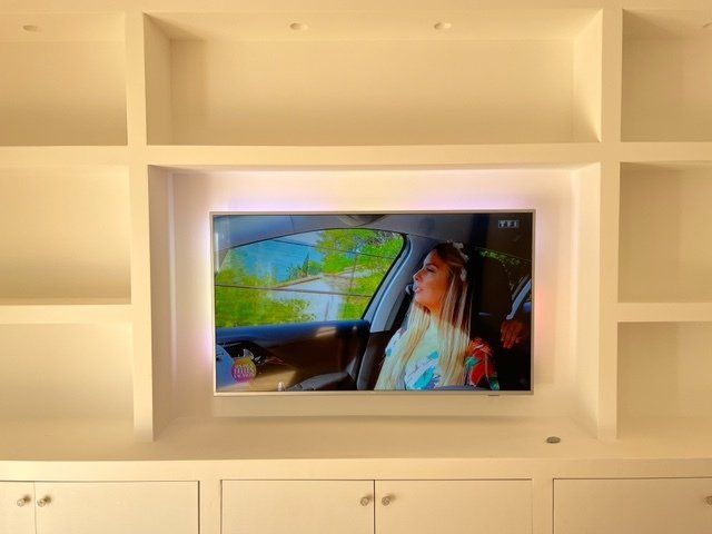 Étagères blanches encastrées avec un téléviseur monté sur lequel figure une personne en voiture. Des lumières LED encadrent l'écran.