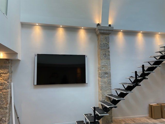 Un grand téléviseur à écran plat monté sur un mur blanc avec un éclairage encastré, des escaliers et une colonne de pierre.