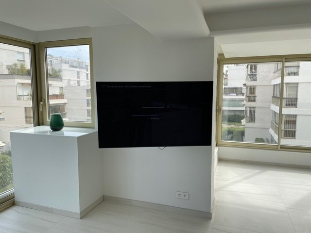 Un salon moderne dispose d'une télévision murale entre deux grandes fenêtres. Un piédestal blanc soutient un vase vert.