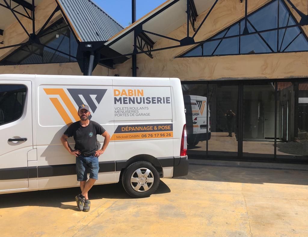 Un homme se tient devant une camionnette blanche portant le logo de Dabin Menuiserie, devant un bâtiment.
