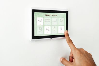 Une main touche un panneau de commande domotique fixé sur un mur blanc. L'écran affiche des icônes.