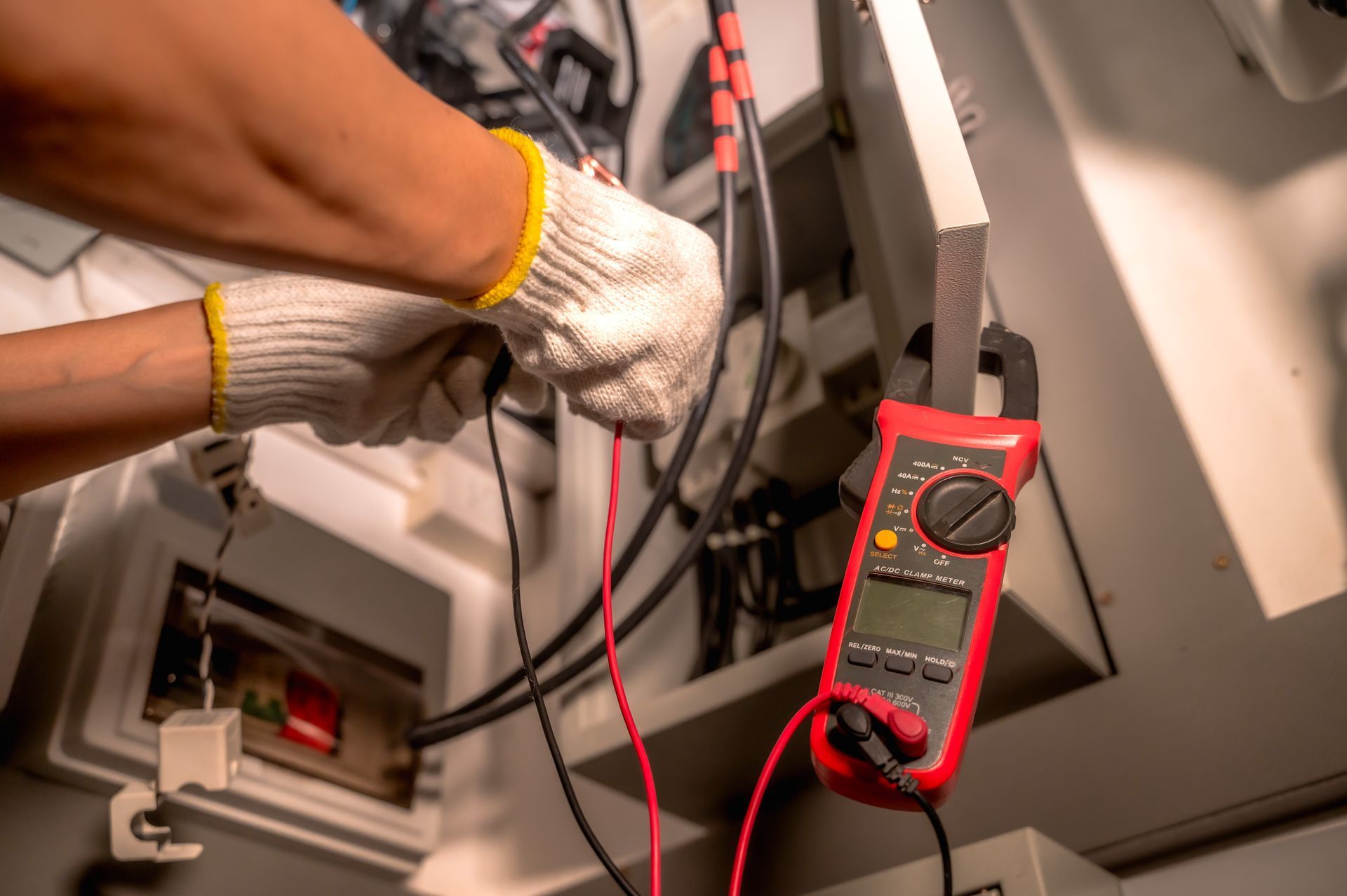 Behandschuhte Hände, die ein rotes Multimeter zum Testen der Verkabelung in einem offenen Verteilerkasten verwenden.
