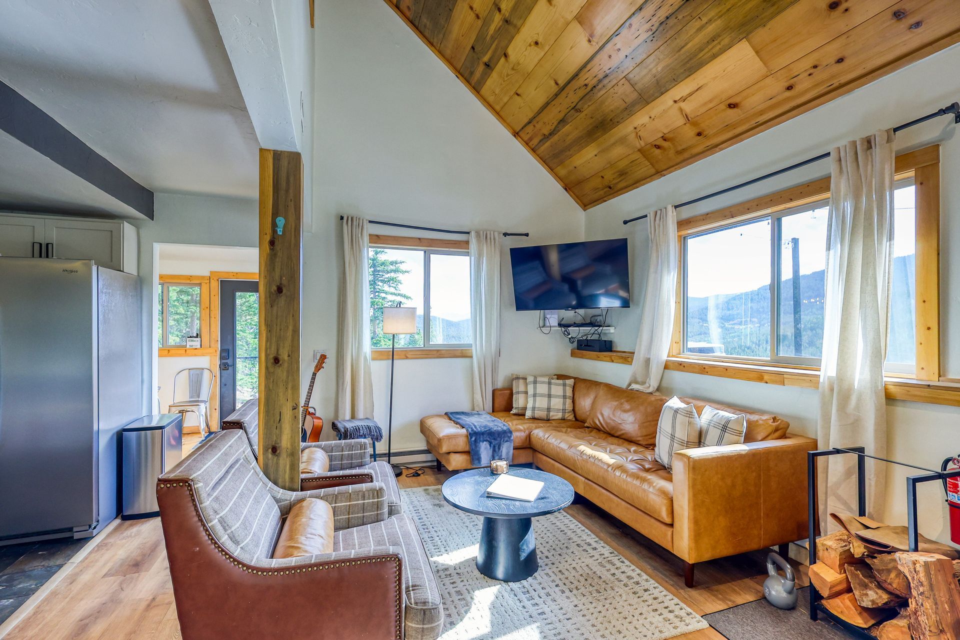 Cozy living room with brown leather sofa, wooden ceiling, windows with mountain view, and fireplace.