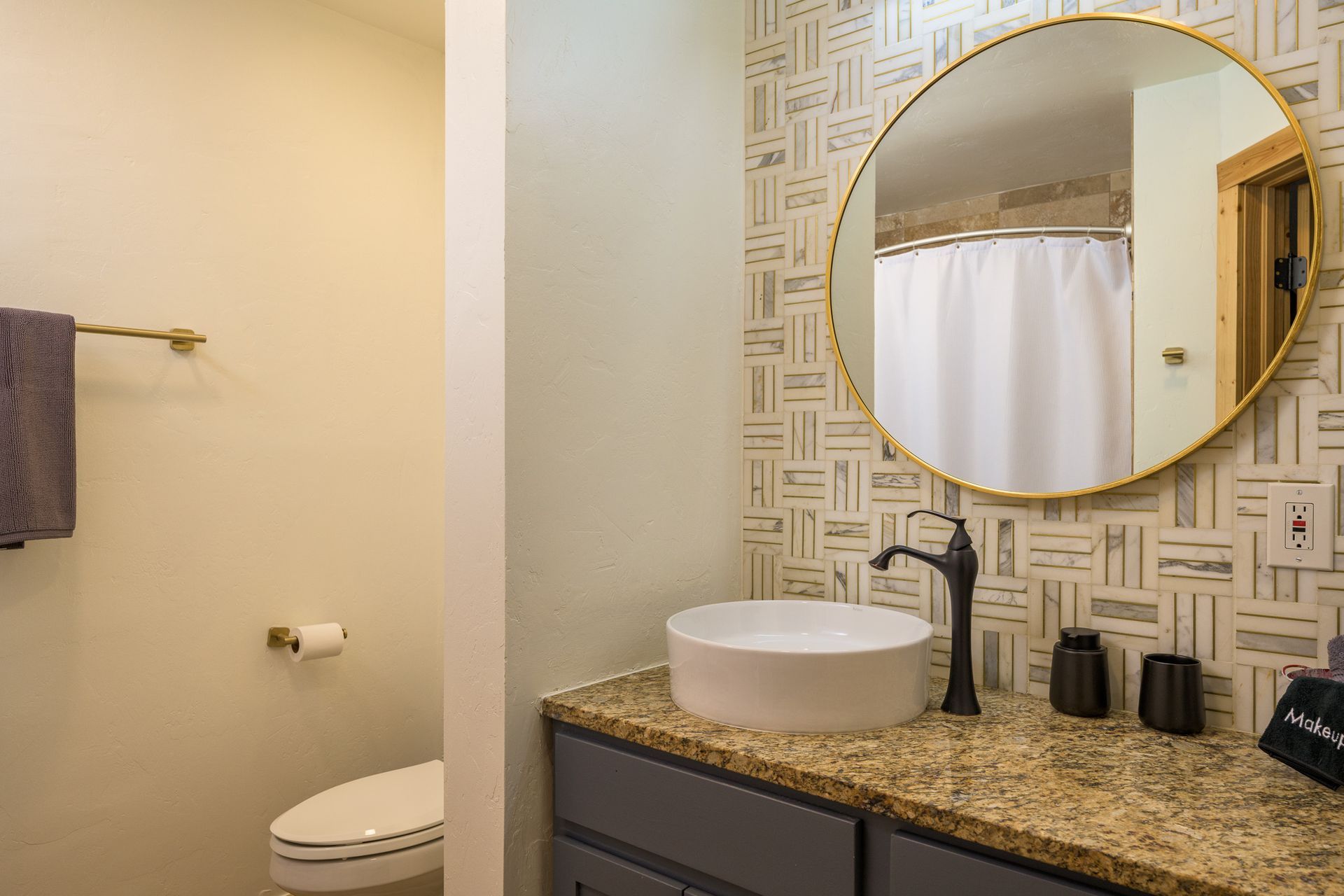 Bathroom with round mirror, vessel sink, patterned wall, toilet, and gold accents.