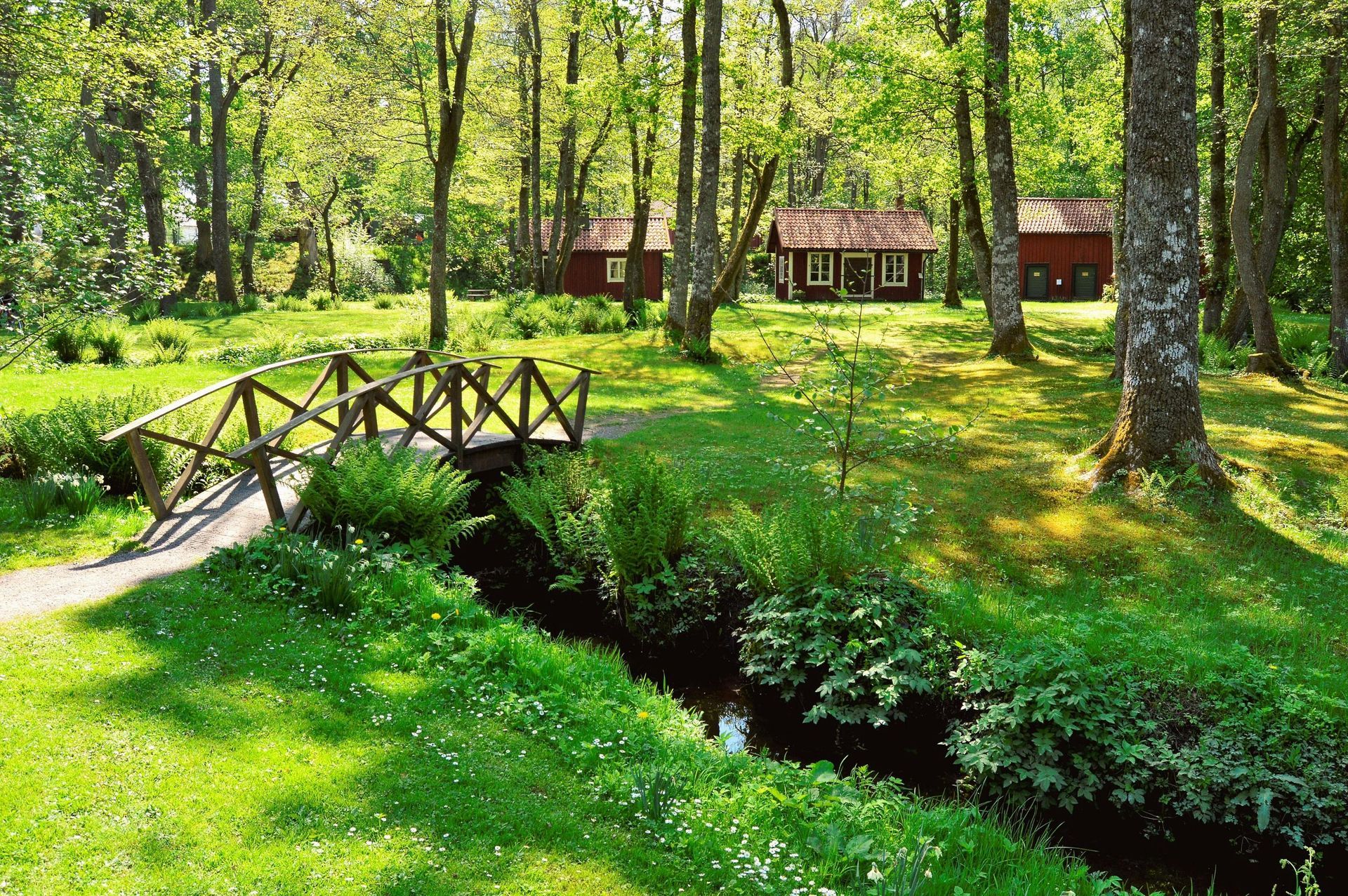 Wooden bridge over stream in lush green park with red cabins.