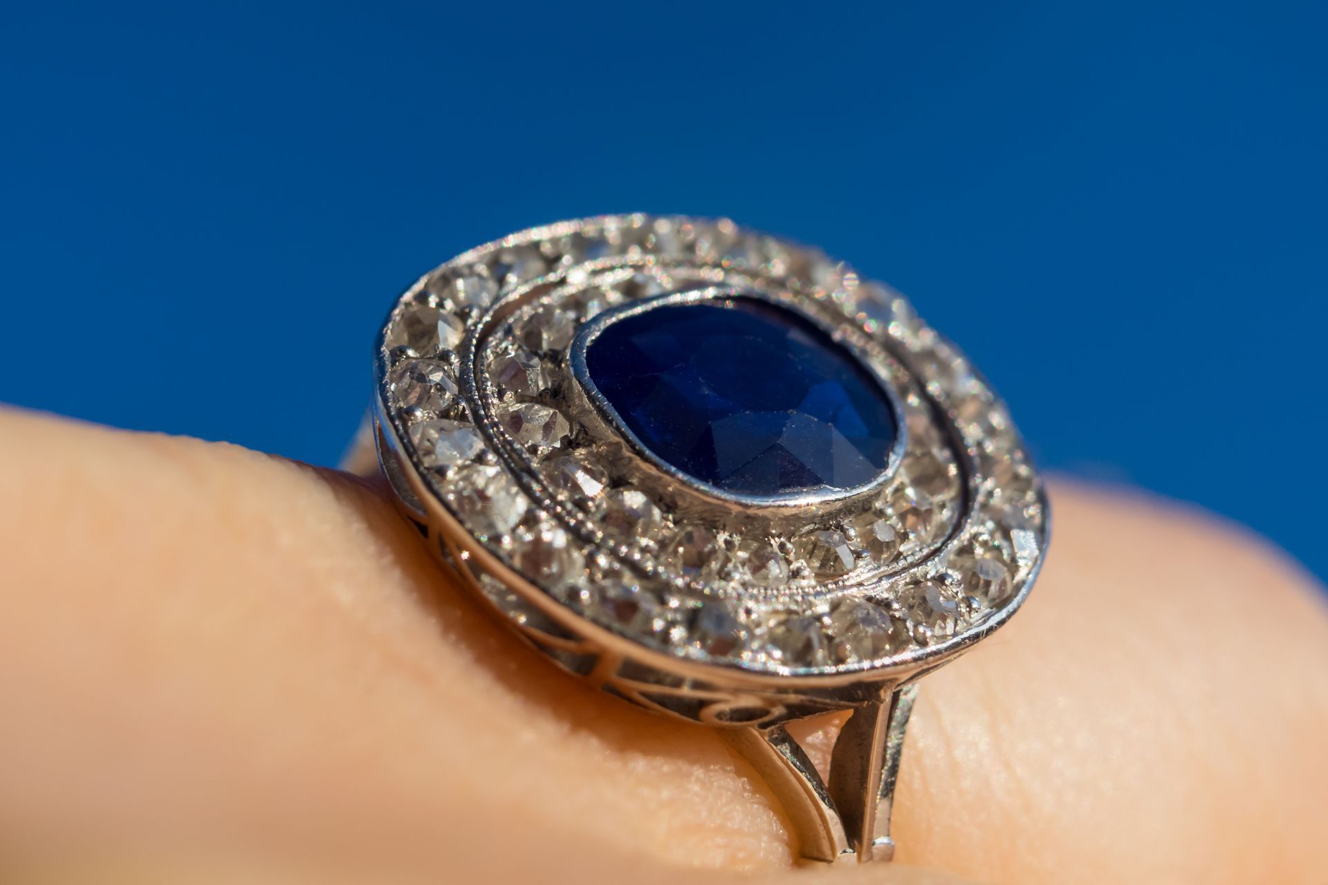 Gros plan sur une bague en argent avec une pierre centrale bleue et des gemmes transparentes tout autour, portée à un doigt.