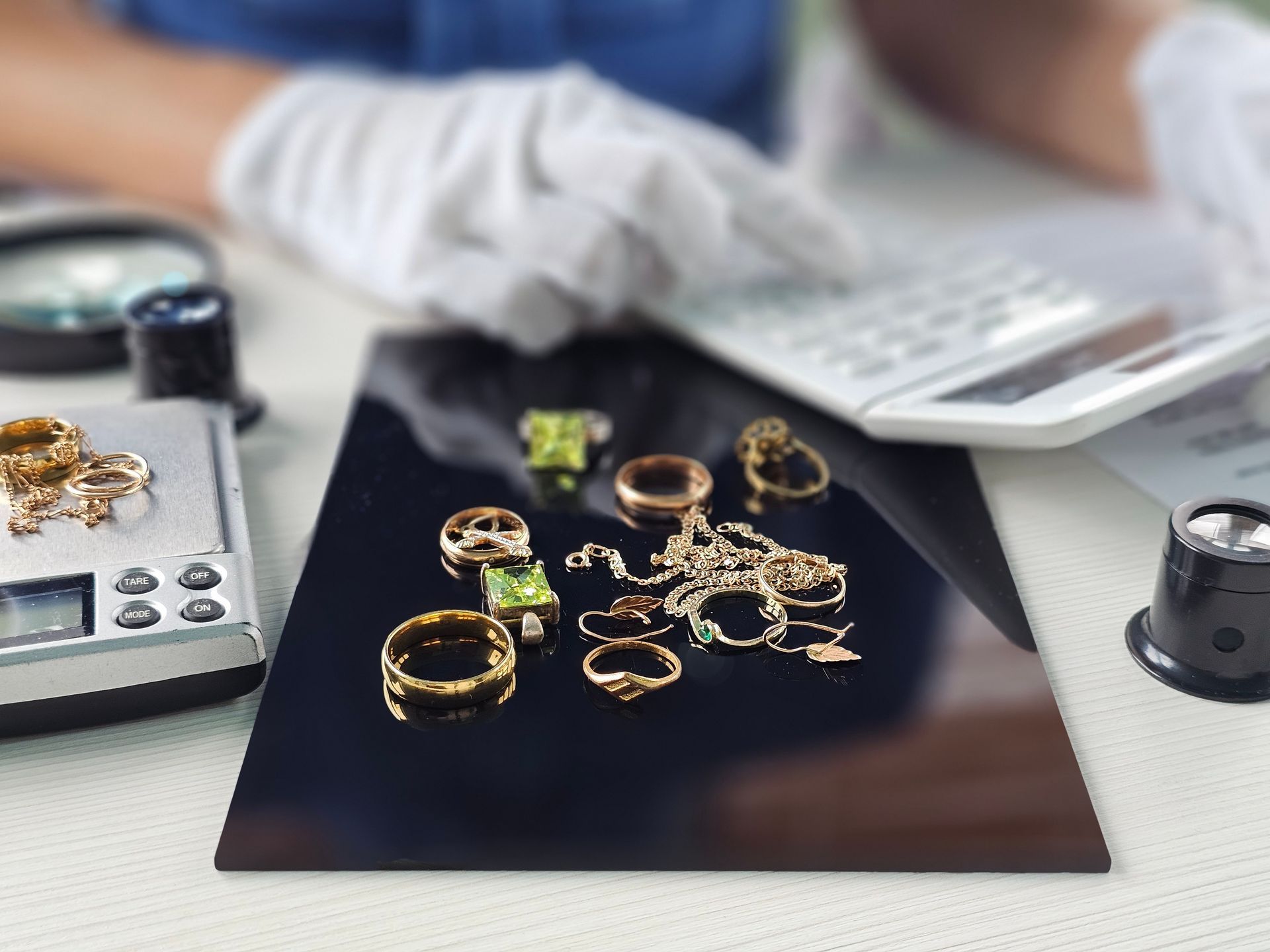 Des mains gantées disposent des bagues et des bijoux en or sur un présentoir noir, des outils à proximité.