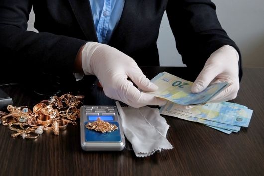 Des mains gantées comptent des billets en euros à côté d'un smartphone, de bijoux et d'un tissu blanc sur une table sombre