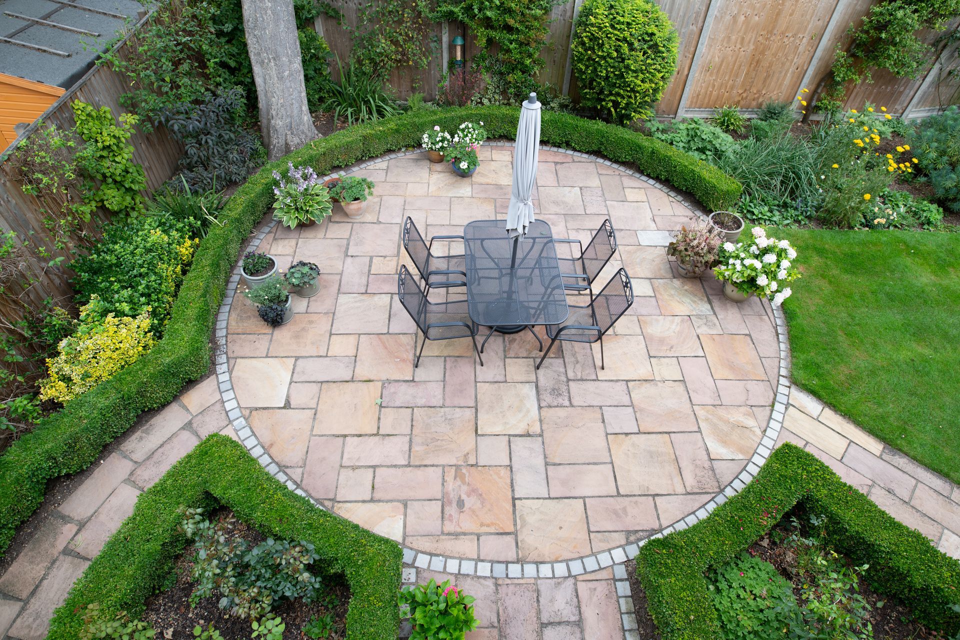 Terrasse circulaire en pierre avec table et chaises, entourée de massifs de fleurs luxuriants et de pelouse.