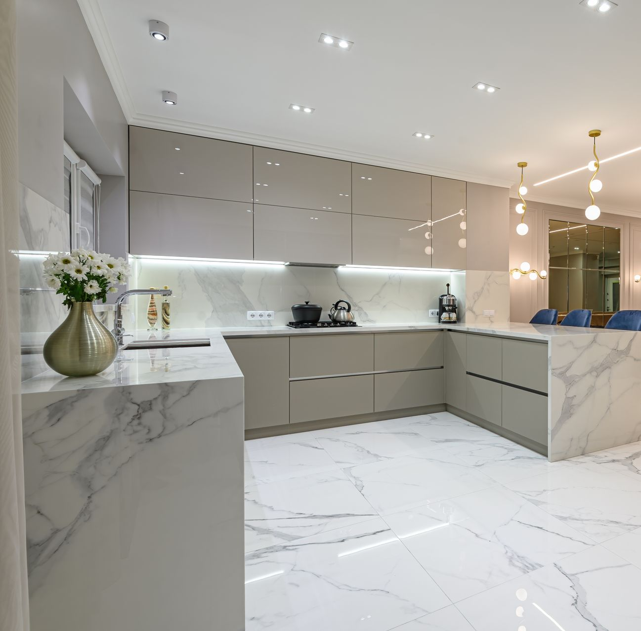Cuisine moderne blanche avec sols et plans de travail en marbre, placards beiges et suspensions lumineuses.