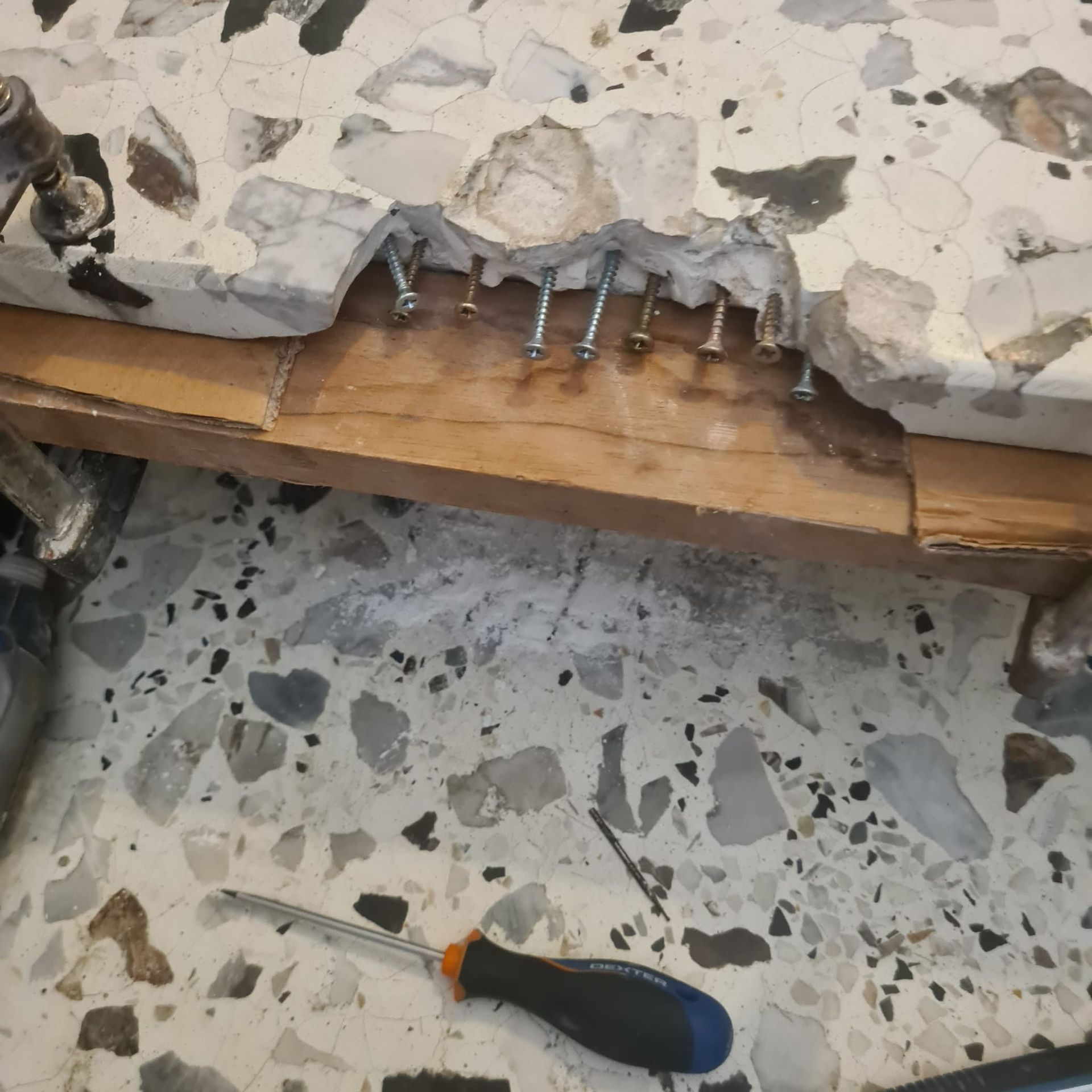 Le sol en terrazzo est endommagé : des vis sont apparentes et un tournevis se trouve sous la fissure.