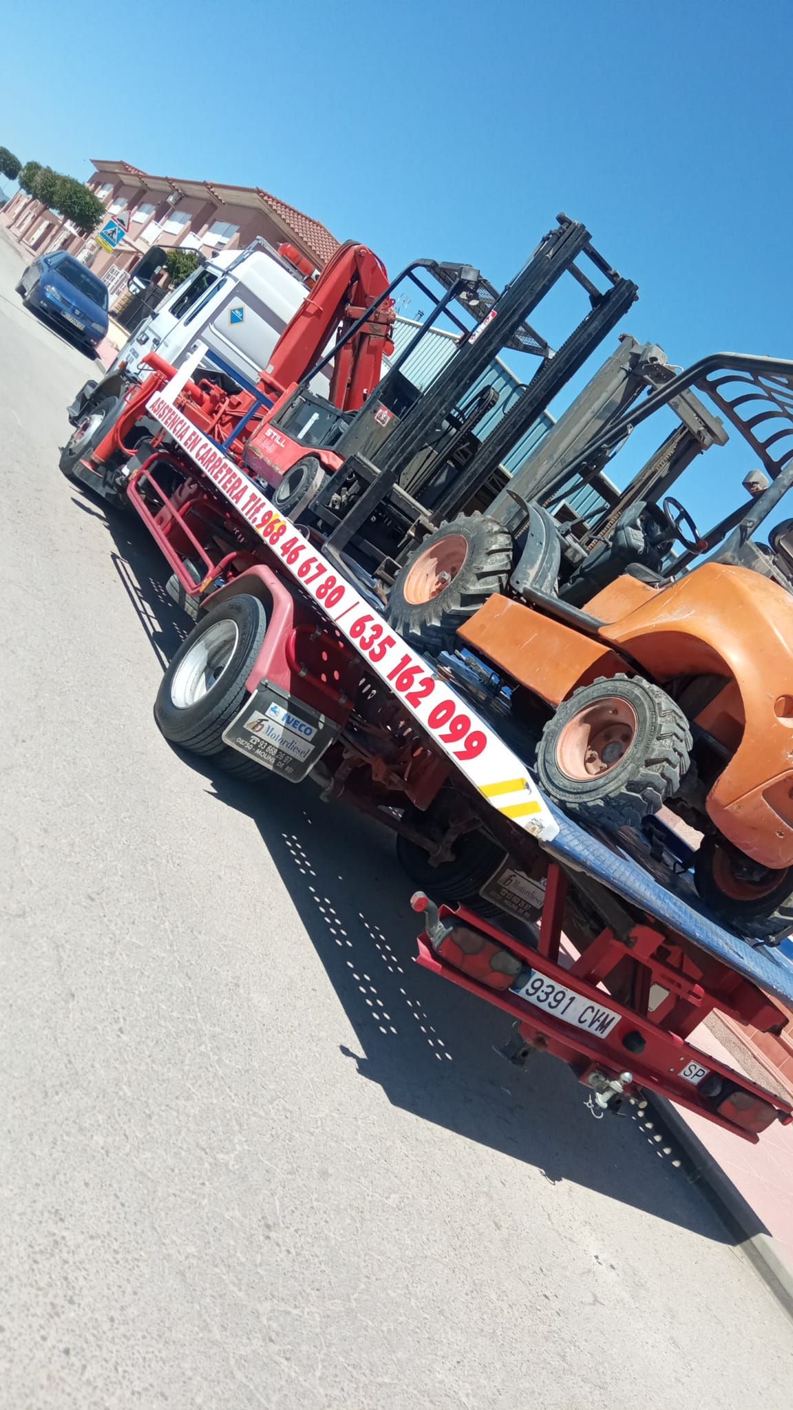Grúa que transporta varias carretillas elevadoras en una calle soleada.