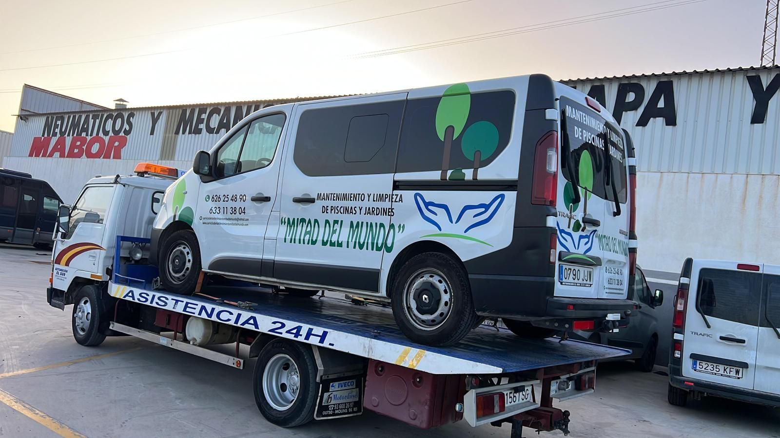 Una furgoneta blanca con detalles verdes sobre una grúa de plataforma. La grúa está estacionada frente a un edificio.
