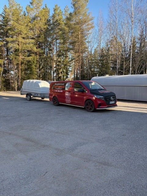 Punainen pakettiauto vetää valkoista katettua perävaunua pysäköintialueella puiden ja suuren lumikinnoksen vieressä.