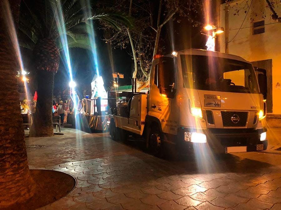 Escena nocturna en una calle con un camión amarillo estacionado junto a una acera iluminada y palmeras, gente a lo lejos.