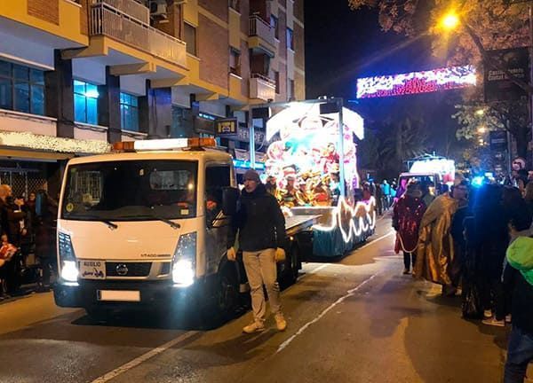 Carroza de desfile nocturno y camión blanco en una calle de la ciudad, con peatones y luces de colores por la noche.