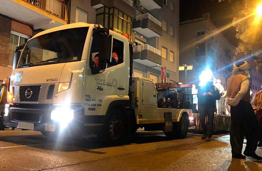 Una grúa de remolque en una calle de la ciudad por la noche, con gente de pie cerca, junto a edificios de apartamentos.