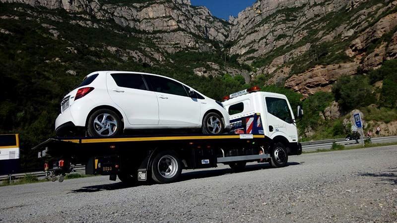 Un coche hatchback blanco sobre una grúa junto a una carretera con colinas rocosas al fondo.
