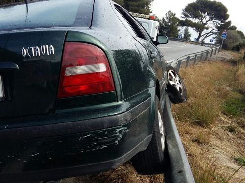 Un coche verde oscuro derrapa fuera de la carretera y queda inclinado sobre el arcén cubierto de hierba junto a la barandilla.