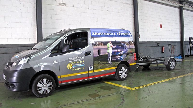 Furgoneta de servicio plateada con remolque aparcada en el interior de un taller, con publicidad en el lateral.