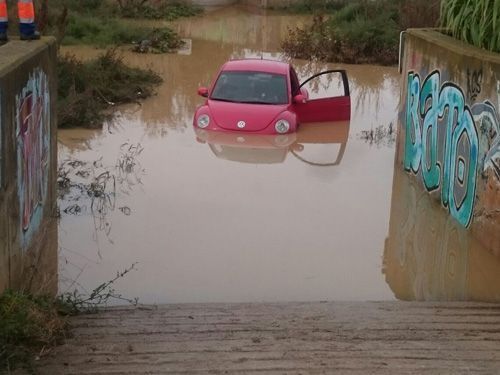 Un coche rosa medio sumergido en el agua de la inundación con la puerta abierta cerca de una pared cubierta de grafitis.