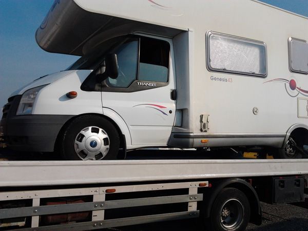 Autocaravana blanca asegurada sobre un camión de plataforma, estacionada al aire libre bajo un cielo nublado.