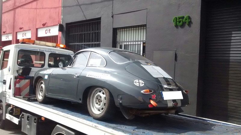 Un coche deportivo clásico gris subido a una grúa frente a un edificio.