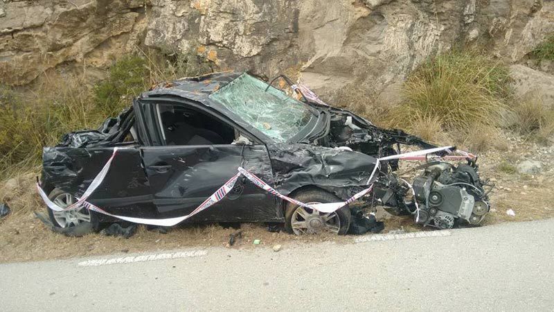 Un coche negro gravemente destrozado quedó aplastado contra un terraplén rocoso al borde de la carretera.