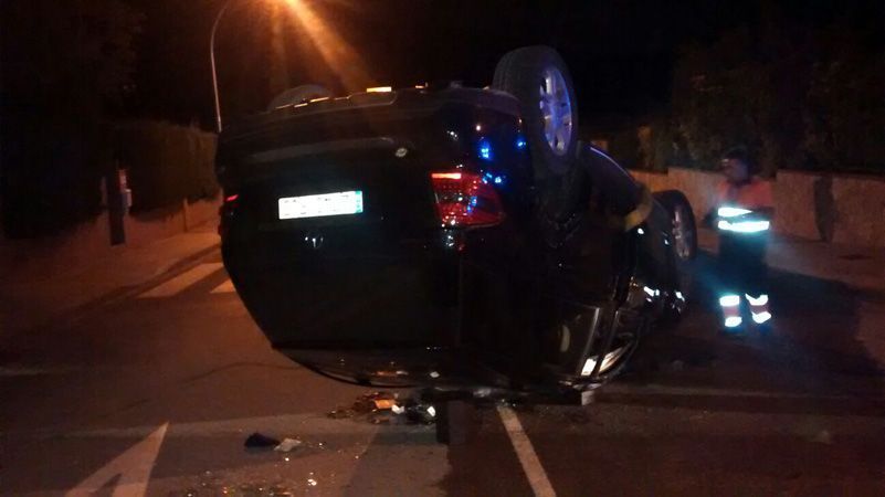 Un SUV negro volcado en una carretera oscura por la noche, con luces intermitentes y gente cerca.