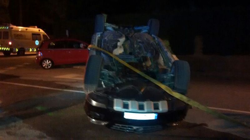 Coche negro gravemente dañado, acordonado con cinta amarilla de precaución en una calle, de noche; vehículo de emergencia cerca.