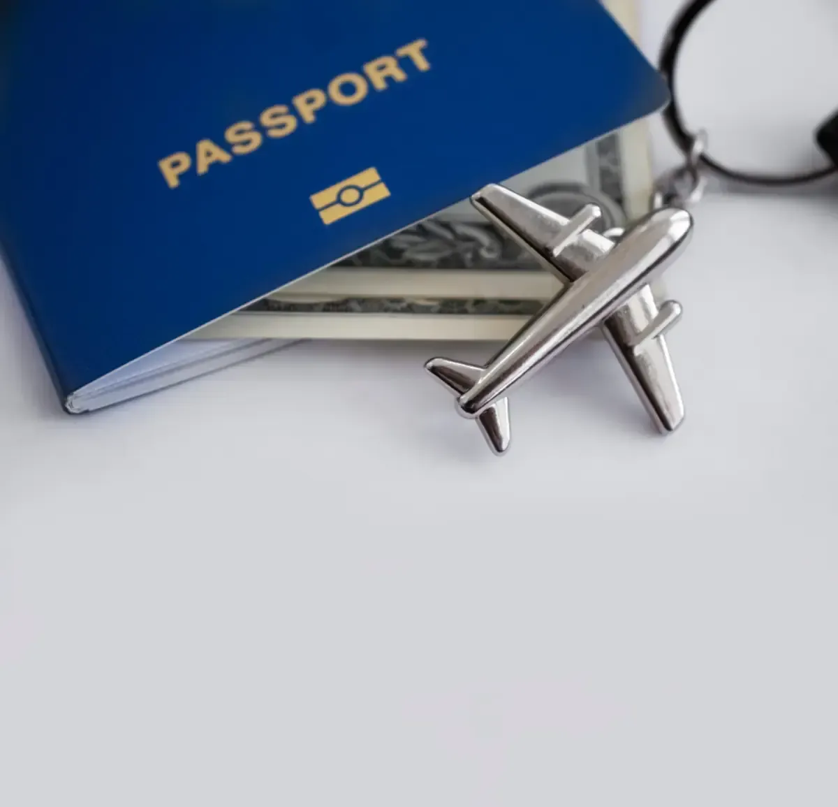 Pasaporte azul con moneda estadounidense y dije de avión sobre superficie blanca.
