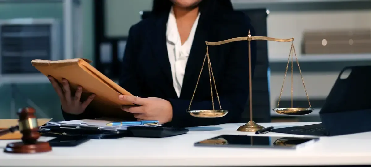 Una persona con traje se sienta en un escritorio, con una balanza de justicia y sosteniendo documentos en un entorno de oficina.