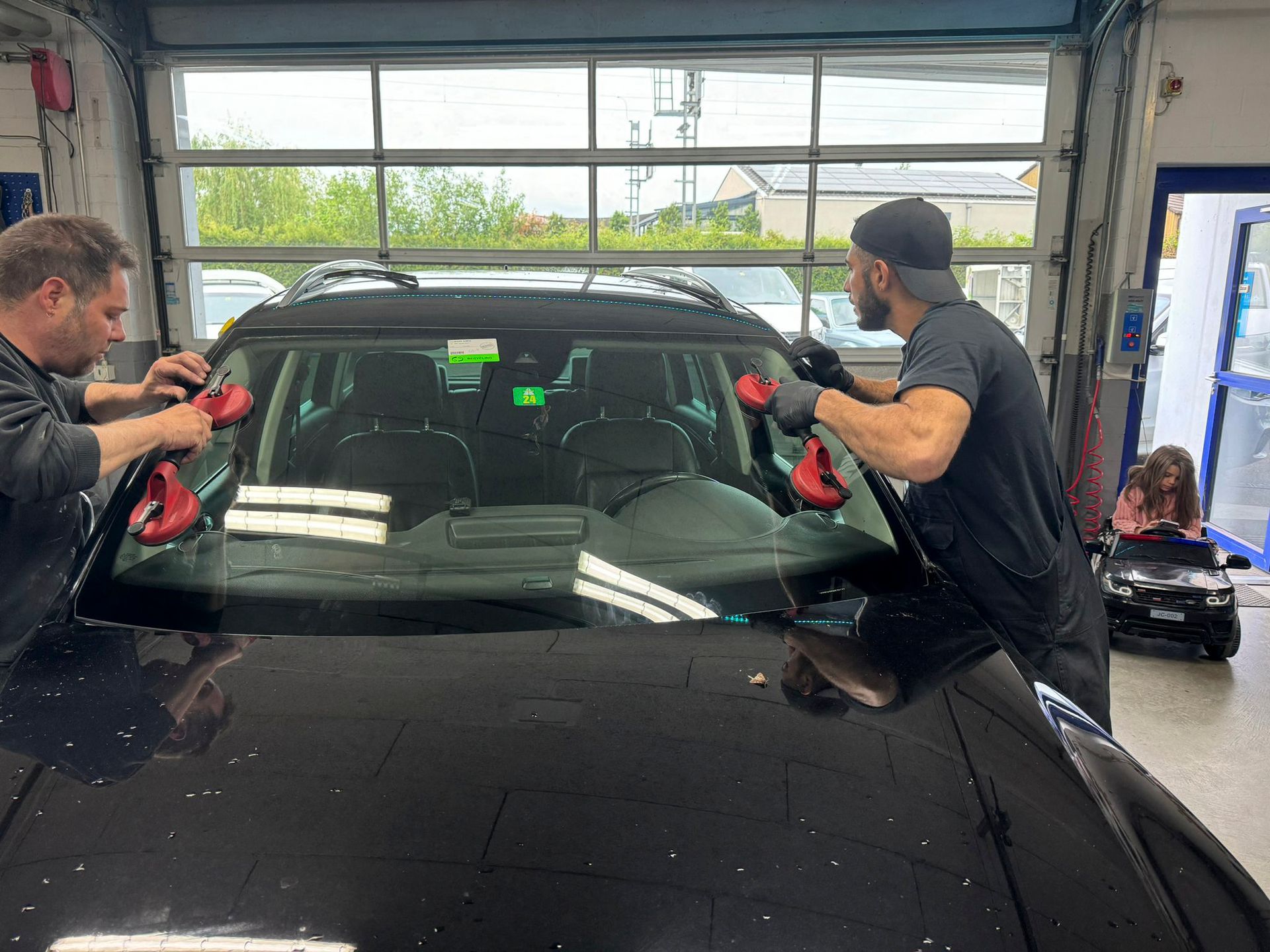 Zwei Männer montieren in einer Garage eine Windschutzscheibe an einem Auto.