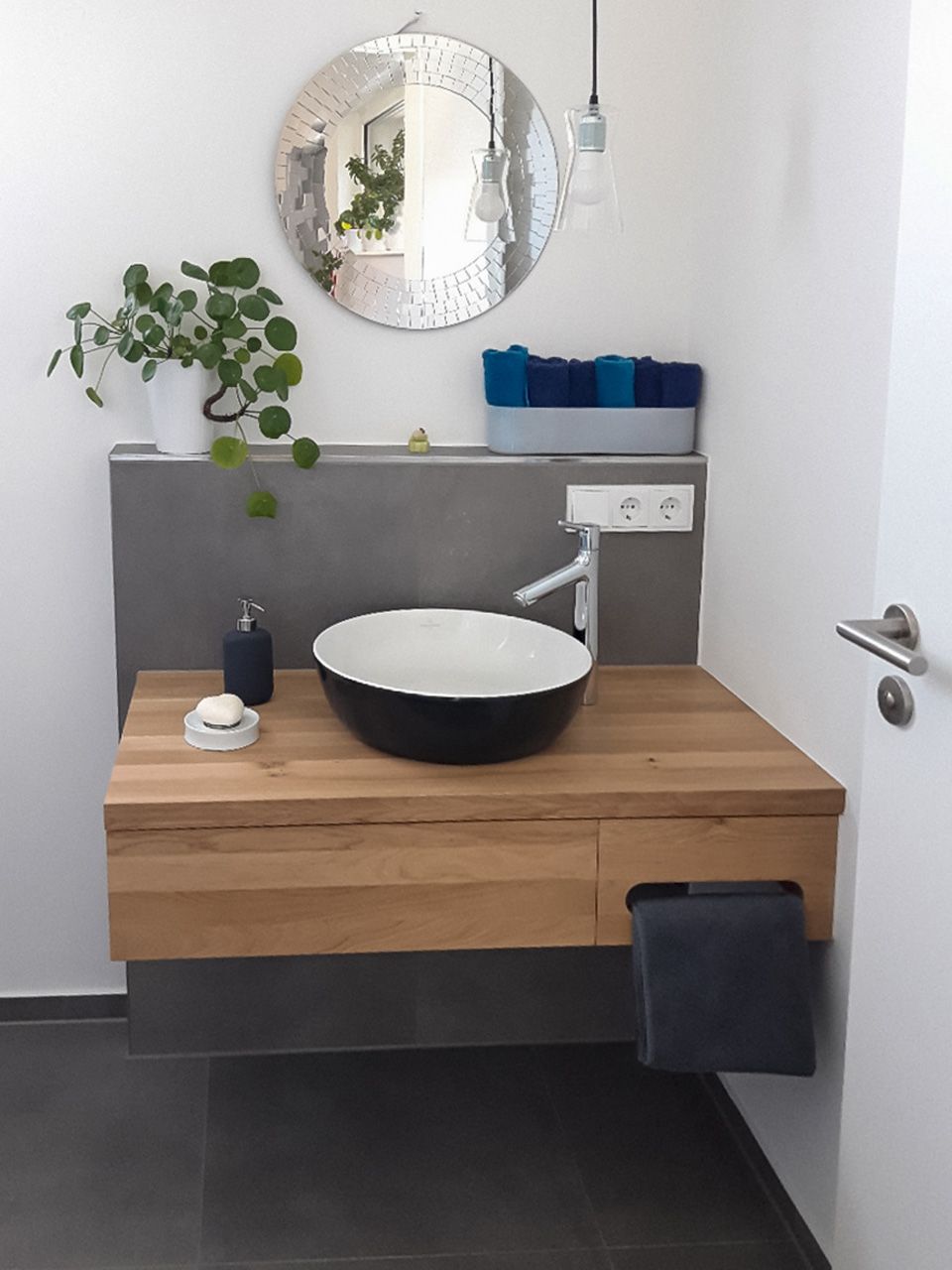 Modernes Badezimmer mit Holzwaschtisch, schwarzem Waschbecken, rundem Spiegel und Topfpflanze.