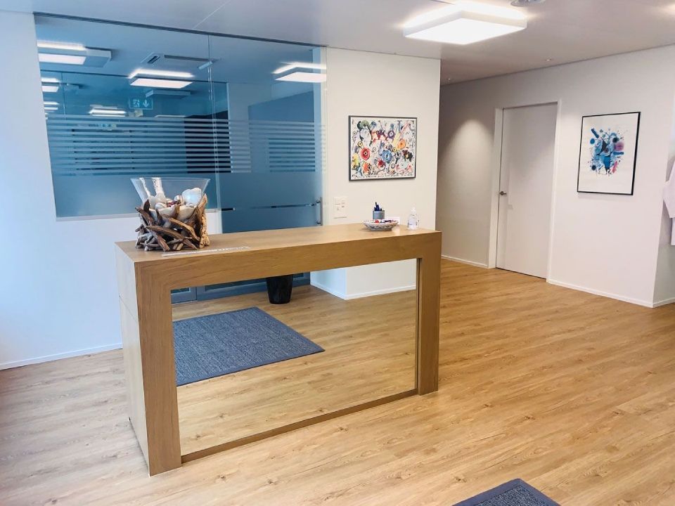 Empfangstresen in einer Klinik mit Kunstwerken an den Wänden und einem Glasfenster. Holzboden und Teppich.